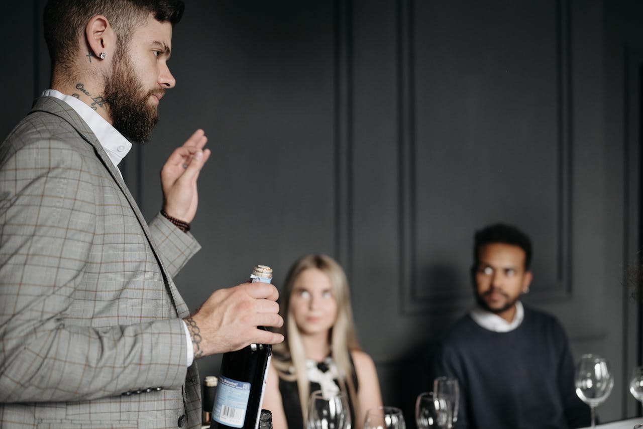 A bearded sommelier in a grey suit presents wine to elegantly dressed guests.